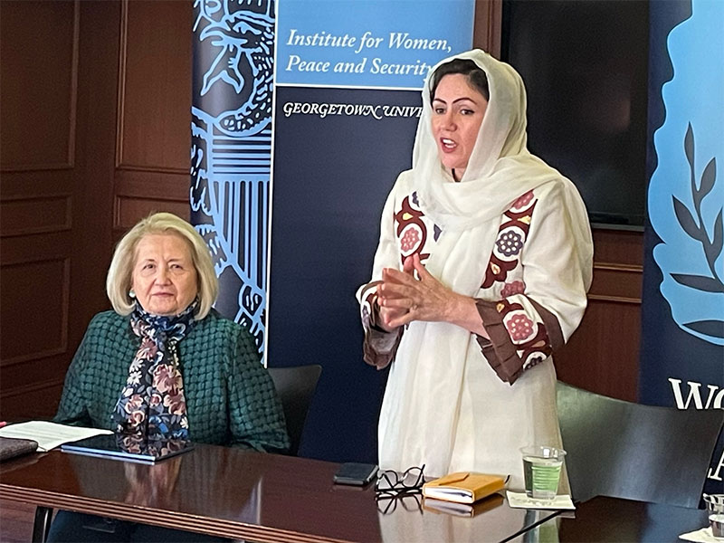 Fawzia Koofia standing up while speaking at at event. Sitting next to her is Ambassador Melanne Verveer.